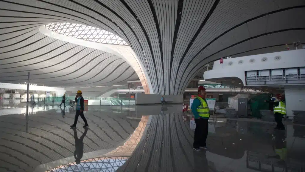 "Interior hall of Beijing Daxing Airport showcasing the steel grid roof structure with C-shaped columns and workers under construction."