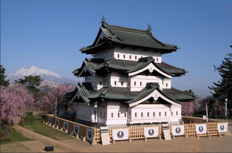 "A traditional Japanese castle showcasing intricate designs with wood and sloped green roofs, set against a backdrop of snow-capped mountains."