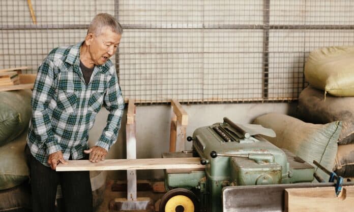 Senior experienced carpenter in plaid shirt putting pine wooden board in thickness planer on workbench