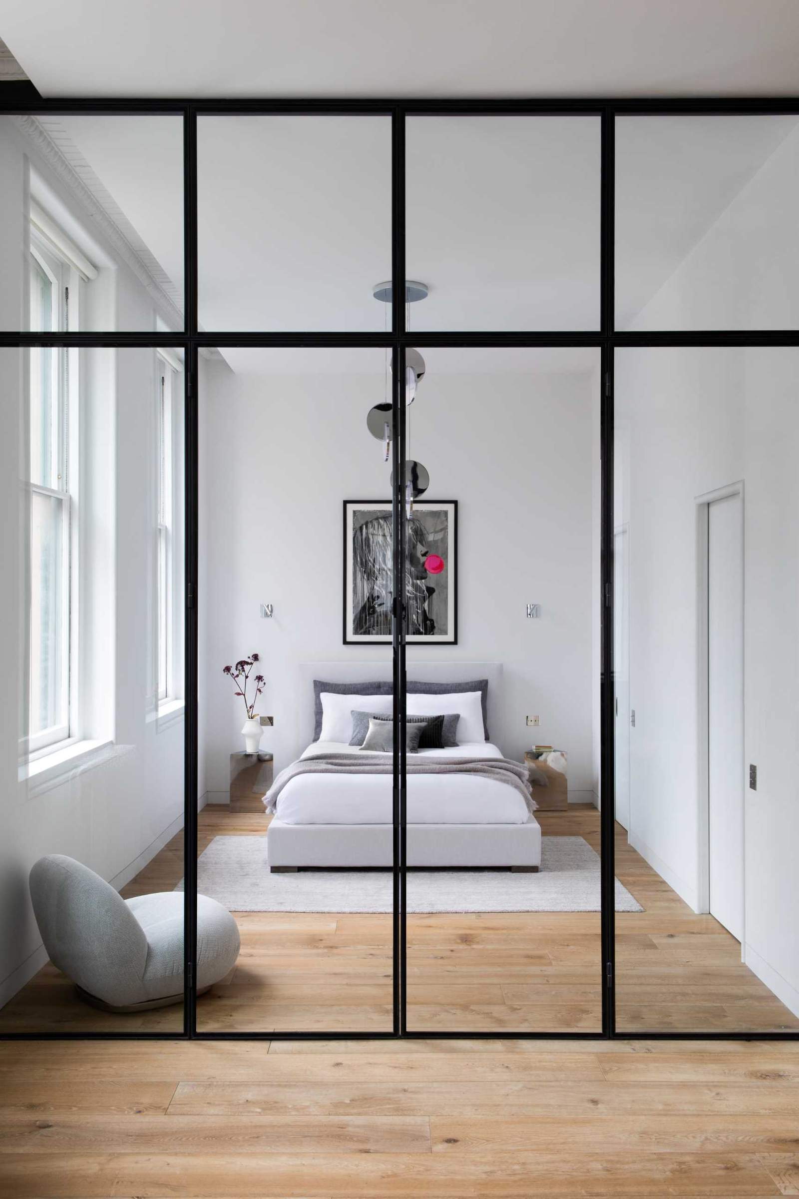 This modern bedroom, located by the windows, includes an Eclipse Chandelier by Lee Broom, a Pacha Lounge Chair by Pierre Paulin, and a Bubble Gum Girl Painting by Hijack.