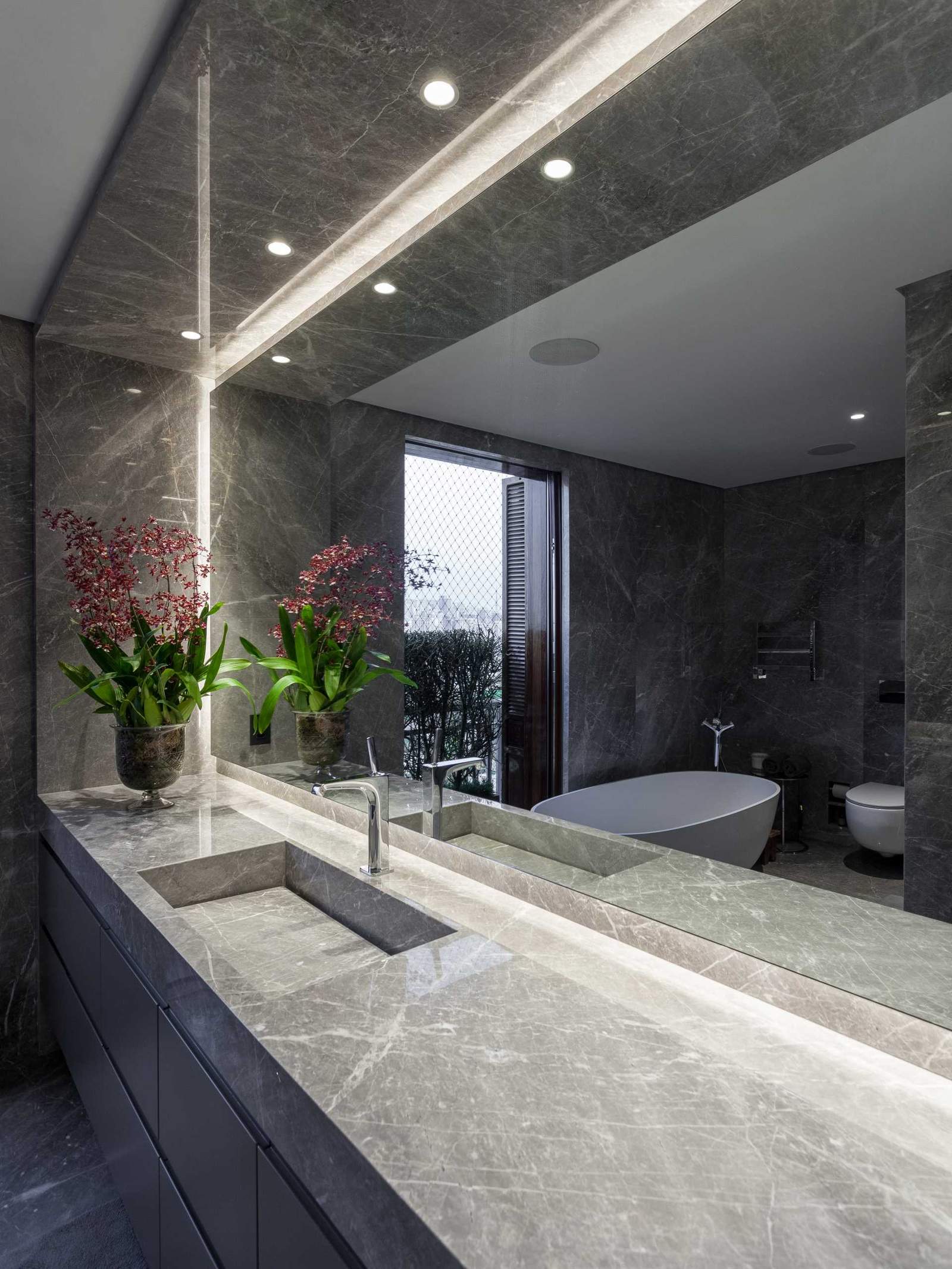 This modern bathroom includes a vanity with built-in sink, a walk-in shower with a bench and shower niche, as well as a freestanding white bathtub by the window.