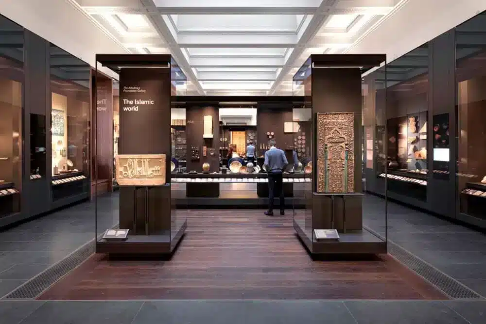 "A museum exhibition space showcasing Islamic artifacts, featuring modern design elements like glass columns and hanging lights that enhance the display's aesthetic appeal."