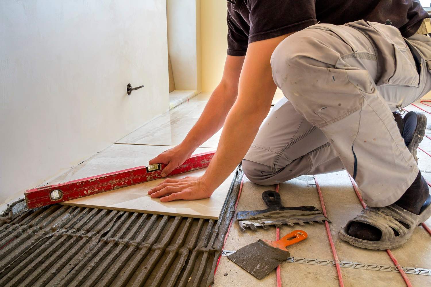 Young worker tiler installing ceramic tiles using lever on cement floor with heating red electrical cable wire system. Home improvement, renovation and construction, comfortable warm home concept.