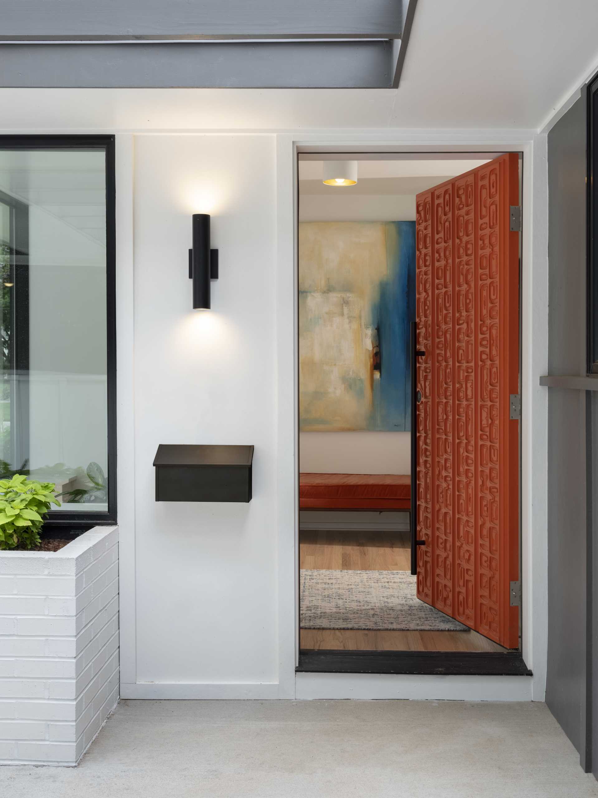 يضيف الباب الأمامي الملون سطوعًا إلى المنزل ويبرز أمام المظهر الخارجي باللونين الرمادي والأبيض، بالإضافة إلى النباتات القريبة.