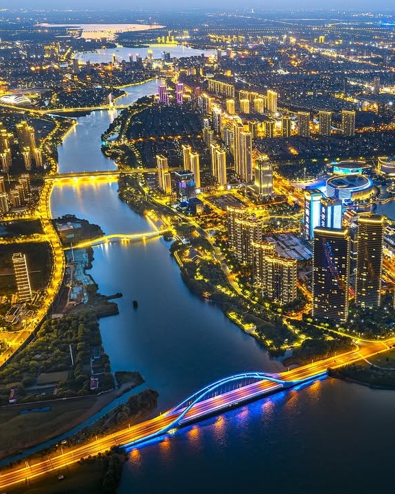 A nighttime view of Wuxi City, showcasing a river flowing through the urban landscape surrounded by modern, brightly lit buildings, as part of the Wuxi Urban Regeneration Competition.