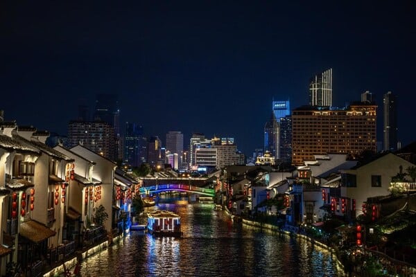 A nighttime view of Wuxi City, showcasing a river flowing through the urban landscape surrounded by modern, brightly lit buildings, as part of the Wuxi Urban Regeneration Competition.