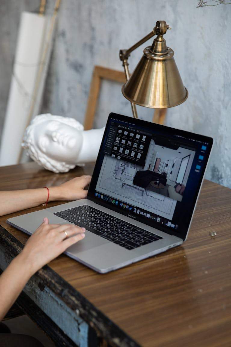 A person working with interior design software on a laptop at a desk with a lamp and statue.