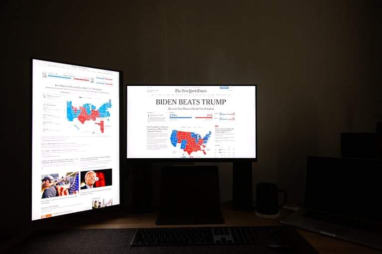 Dual computer screens in a dark room display election results indicating Biden's victory over Trump.