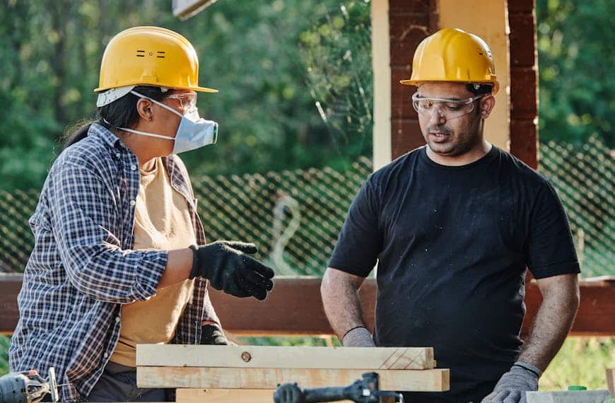 Two construction workers discussing plans at an outdoor site, wearing protective gear.
