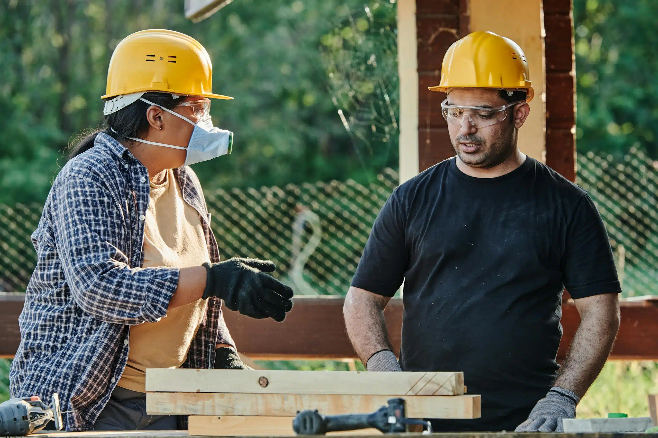 Two construction workers discussing plans at an outdoor site, wearing protective gear.