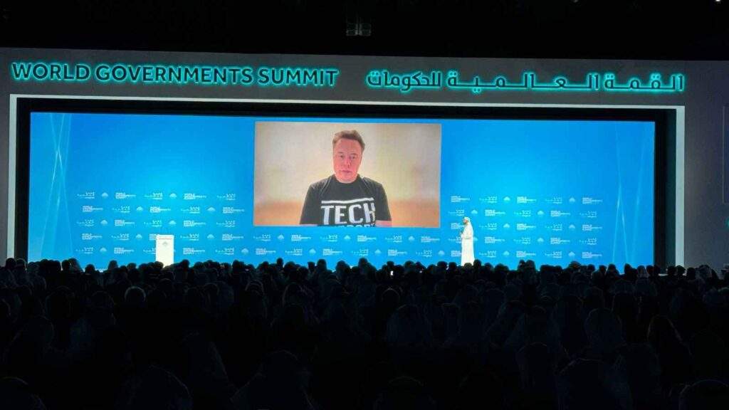 A wide-angle view of Elon Musk speaking via video call at the World Governments Summit 2025 in Dubai, with Omar Sultan Al Olama on stage, addressing the audience about the Dubai Loop transportation project.