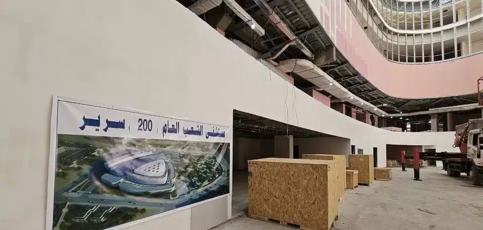 صورة داخلية لمستشفى تحت الإنشاء تُظهر أعمال البناء والتطوير، مع لافتة تحمل تصميم المستشفى النهائي