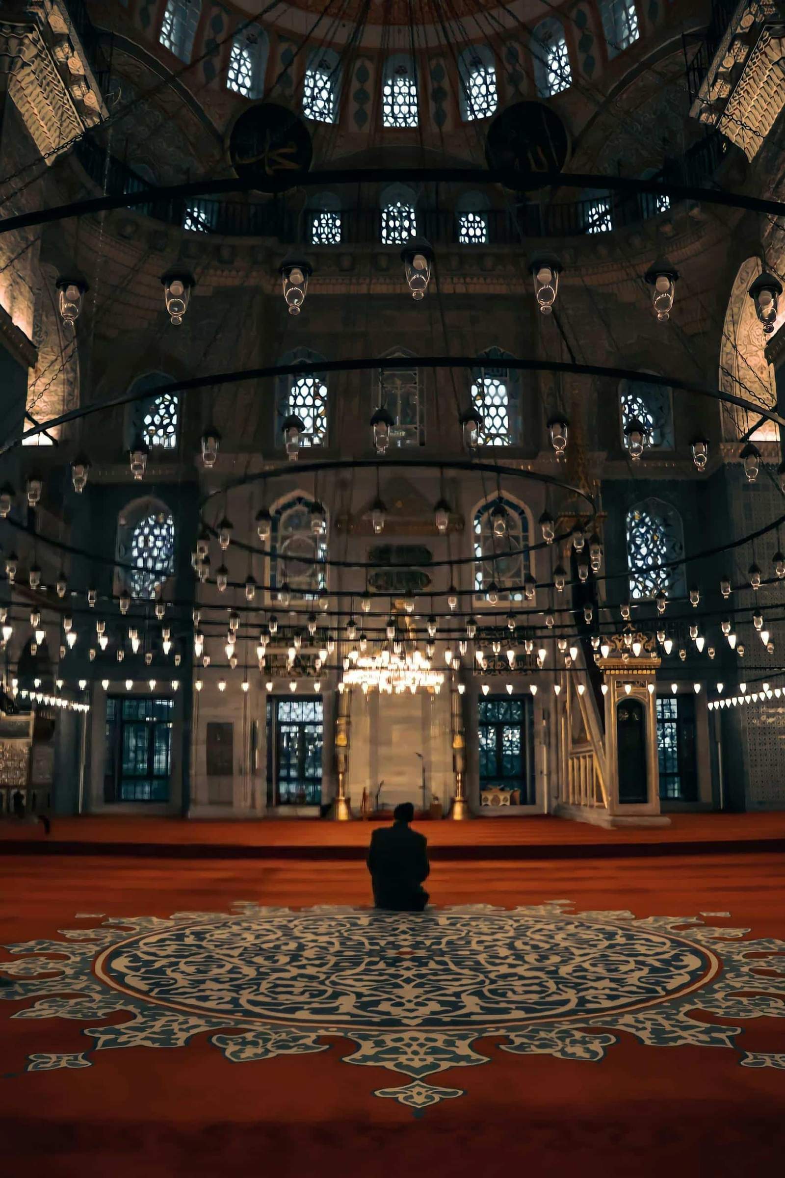 A person praying in a beautifully lit, ornate Ottoman-style mosque interior in Istanbul, Turkey.