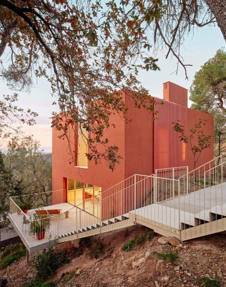 "Modern sustainable house on a steep hillside with natural tones, featuring an outdoor terrace and cantilevered stairs connecting levels."