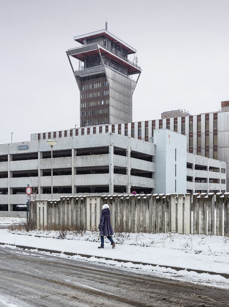 A woman walks past a multi-story concrete parking garage, depicting daily life in post-war suburbs.