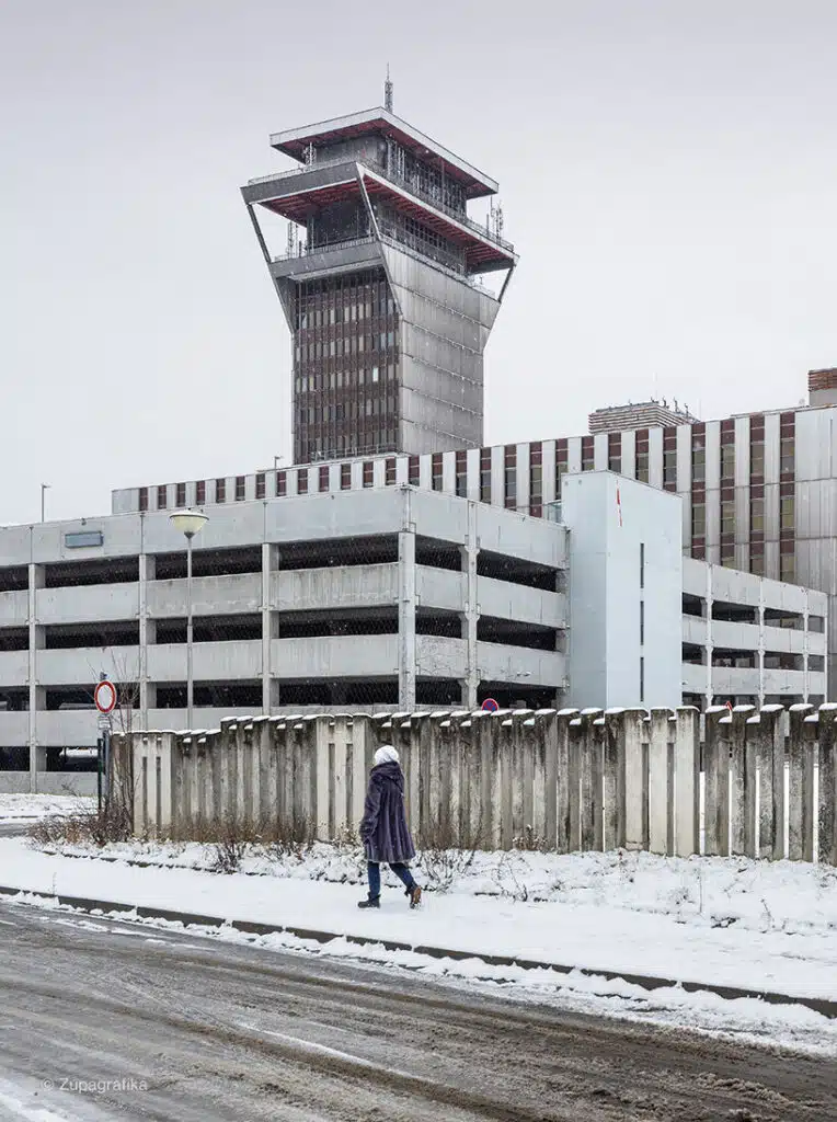 A woman walks past a multi-story concrete parking garage, depicting daily life in post-war suburbs.