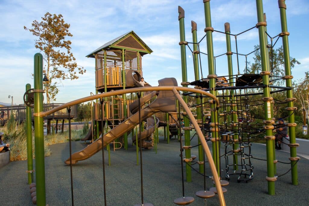 A playground in a public park featuring slides, climbing nets, and elevated play towers, surrounded by trees and clear skies.