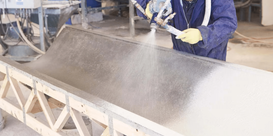 A worker wearing protective clothing and gloves is spraying a mixture of Glass Fiber Reinforced Concrete (GRC) onto a wooden mold using a water-based spray system. The process illustrates the manufacturing stage of GRC in a factory setting
