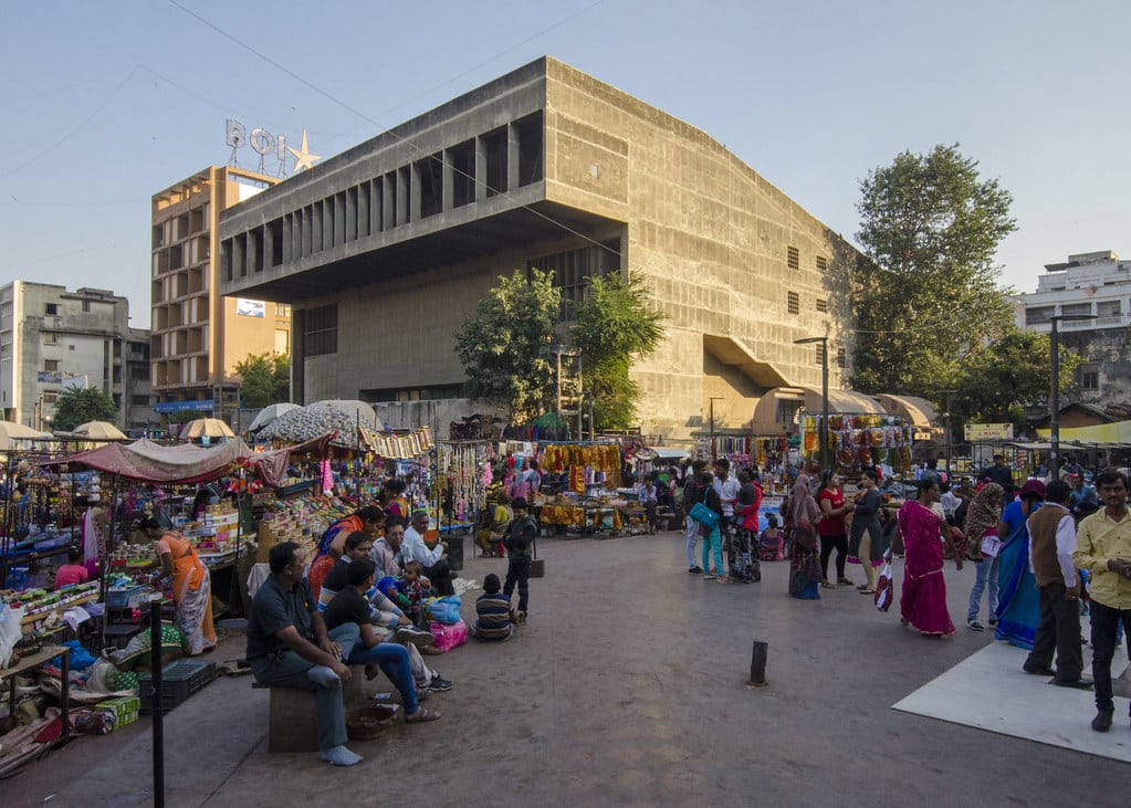 A bustling street market with people shopping and sitting in an open area in front of a modern building with flat roofs and concrete pillars.