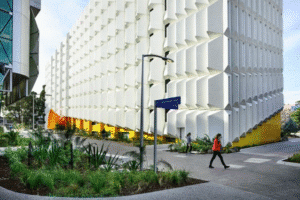 Modern building facade covered with Glass Fiber Reinforced Concrete (GRC) panels in a honeycomb-like geometric design, featuring yellow decorative accents at the base. The area is clean and professionally designed, with pedestrians crossing the street.