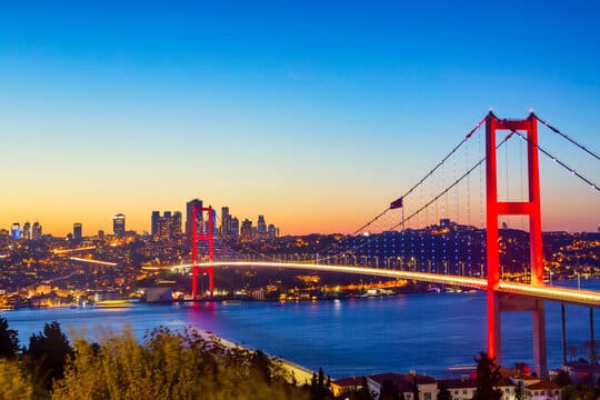 A large suspension bridge with bright red towers spans a wide river, connecting two urban areas. The sky in the background shows warm hues of dusk, with city lights glowing against the darkening sky