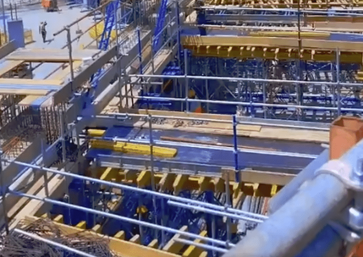 Construction workers are seen working on the upper tier of a stadium, with scaffolding and formwork systems in place. The site is equipped with safety railings and structural supports, indicating an organized and safe construction environment.