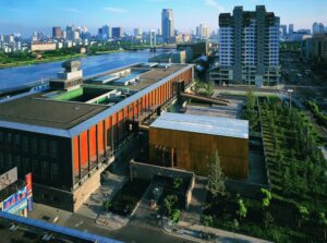An aerial view of a modern architectural complex with a mix of flat and sloped roofs, featuring vibrant orange and wooden facades. The building is surrounded by greenery and urban elements, including a river in the background and high-rise buildings in the distance.