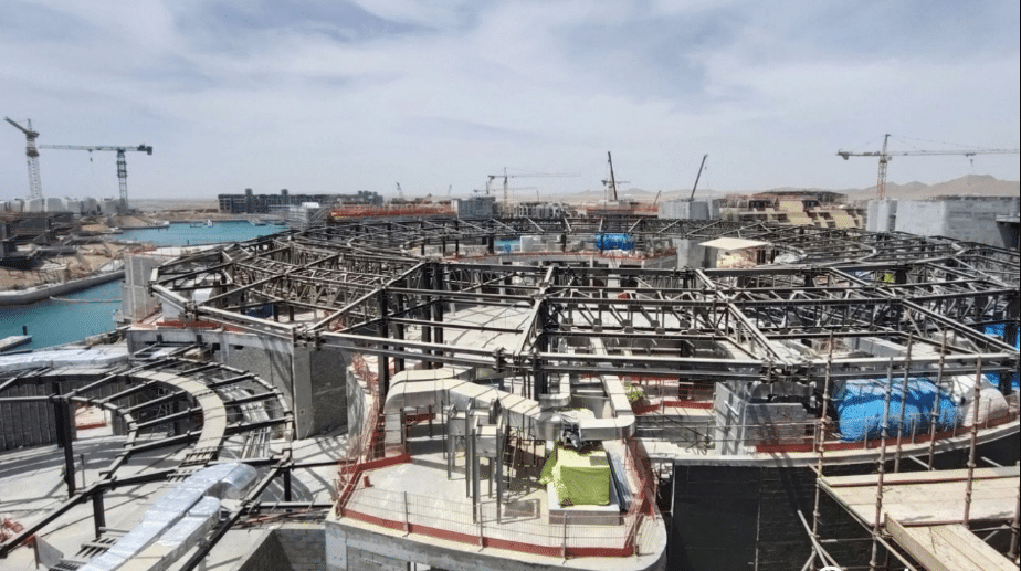 A sweeping view of a construction site along the Red Sea coastline, showcasing intricate steel frameworks and circular concrete structures that blend seamlessly with the natural surroundings. The site is bustling with activity, featuring cranes and construction materials, all set against a backdrop of clear blue skies and calm waters