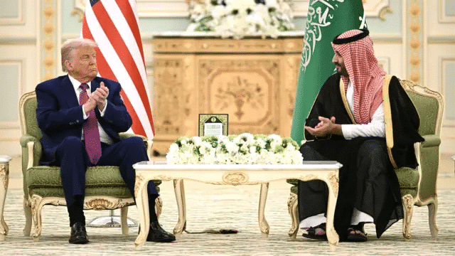 A formal meeting between two world leaders seated in an opulent room, both dressed in formal attire. Between them is a small table adorned with white flowers, with the flags of the United States and Saudi Arabia visible in the background.