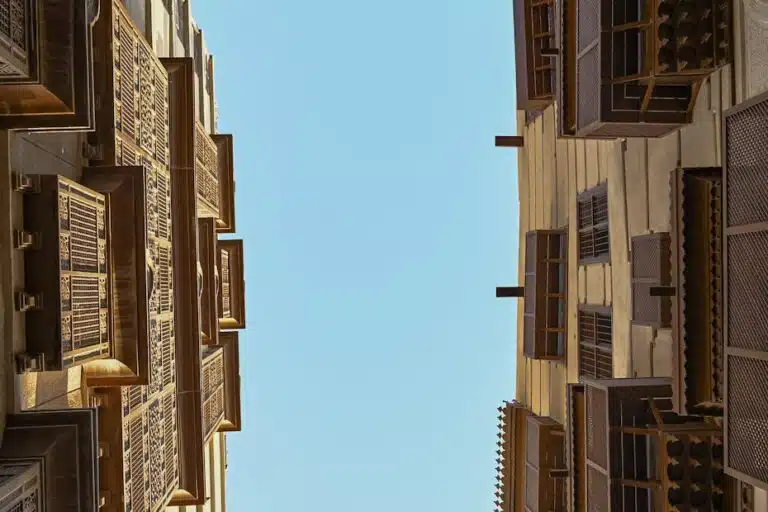 A close-up view of traditional Islamic architecture, showcasing intricate wooden screens and geometric patterns. The structures are adorned with detailed carvings and layered designs, allowing sunlight to filter through in a mesmerizing dance of light and shadow. The warm tones of the wood contrast beautifully against the clear blue sky, emphasizing the timeless beauty and functionality of these architectural elements.
