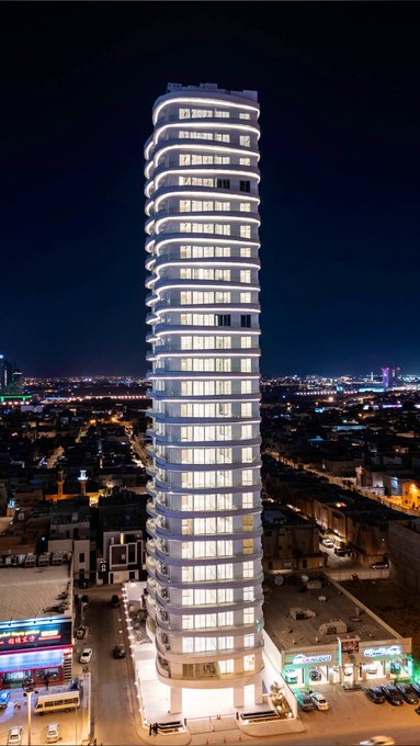 At night, The Oval Residential Tower is illuminated, highlighting its elegant curves and glassy exterior, creating a striking silhouette against the city lights of Riyadh.