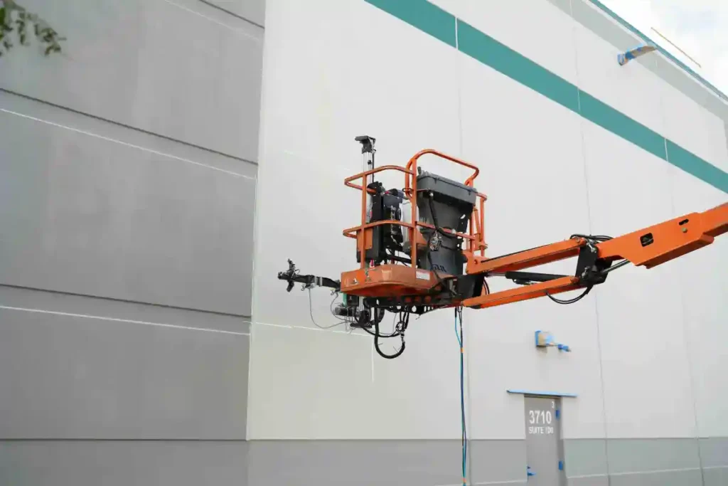  An orange industrial lift raises a machine or device to a high elevation on the side of a building, showcasing the use of technology for challenging tasks.