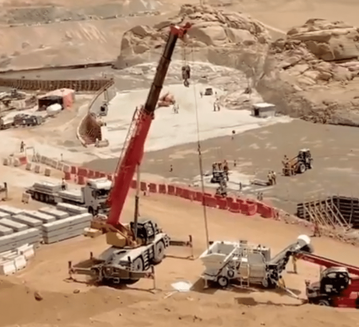 Aerial view of a large construction site in a desert area, featuring heavy machinery such as a red crane and trucks working to prepare the ground. Workers in safety gear are coordinating with equipment to carry out infrastructure work.