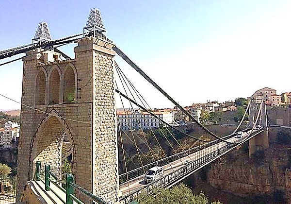 A traditional suspension bridge with a stone structure and decorative arches spans a small river. The bridge carries a single vehicle and is surrounded by a serene natural landscape with trees and residential buildings in the background.