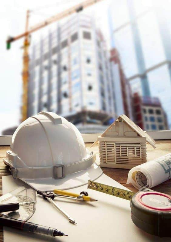 A desk filled with engineering tools, including a hard hat, measuring tape, pencil, and a small house model, with a building under construction in the background