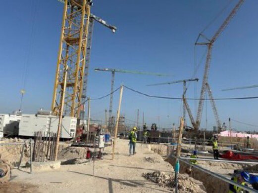 A construction site showing workers in safety harnesses and helmets, with large cranes and other machinery visible in the background.