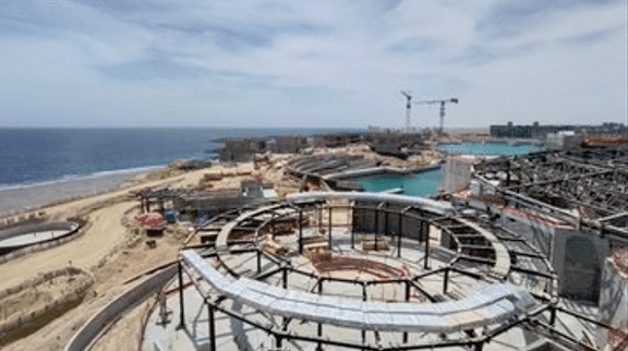 An aerial view of the AMAALA construction site, highlighting the organic, flowing design of the structures. The site features curved steel frameworks and modern architectural elements that seamlessly integrate with the natural landscape of the Red Sea coastline. The surrounding area is a mix of desert sand and calm waters, with cranes and construction materials visible in the background.