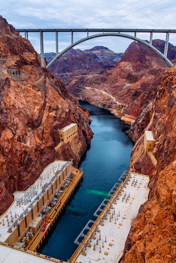 The massive Hoover Dam, suspended between towering red rock cliffs, with the vibrant blue Colorado River flowing beneath it.