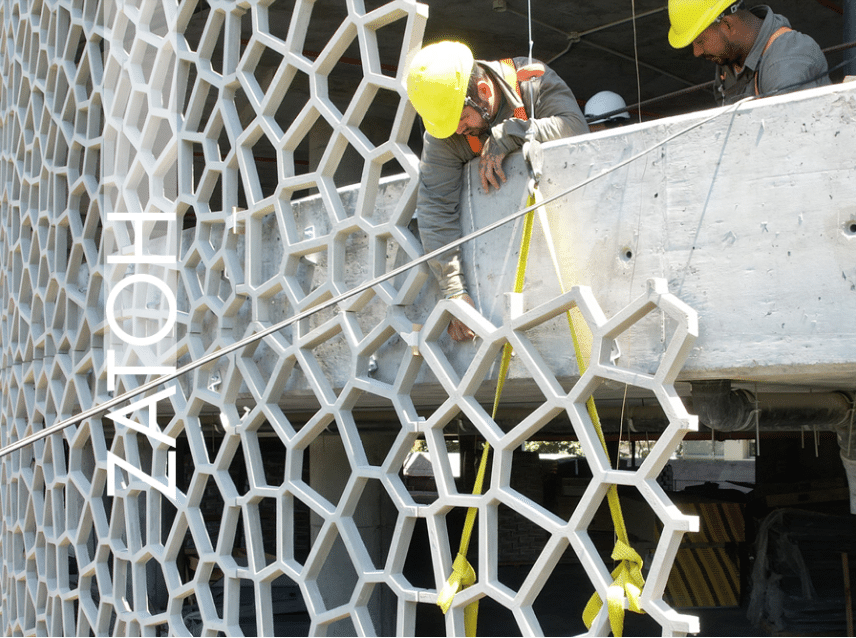 Construction workers wearing yellow safety helmets are installing Glass Fiber Reinforced Concrete (GRC) panels with a three-dimensional, honeycomb-like geometric pattern. The facade under construction showcases a unique architectural design, with the word ZAOH written on the surface.