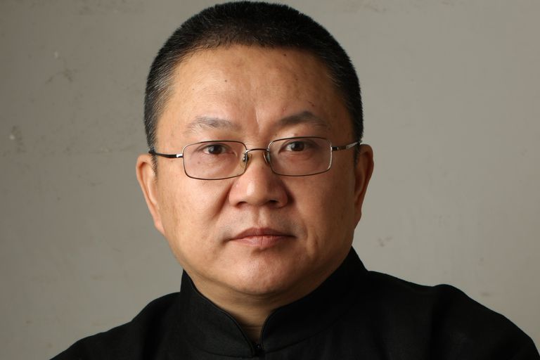A man with short dark hair and glasses, wearing a black traditional-style shirt, is seen in a close-up portrait. His expression is calm and thoughtful, with a slight smile, against a plain, neutral background.