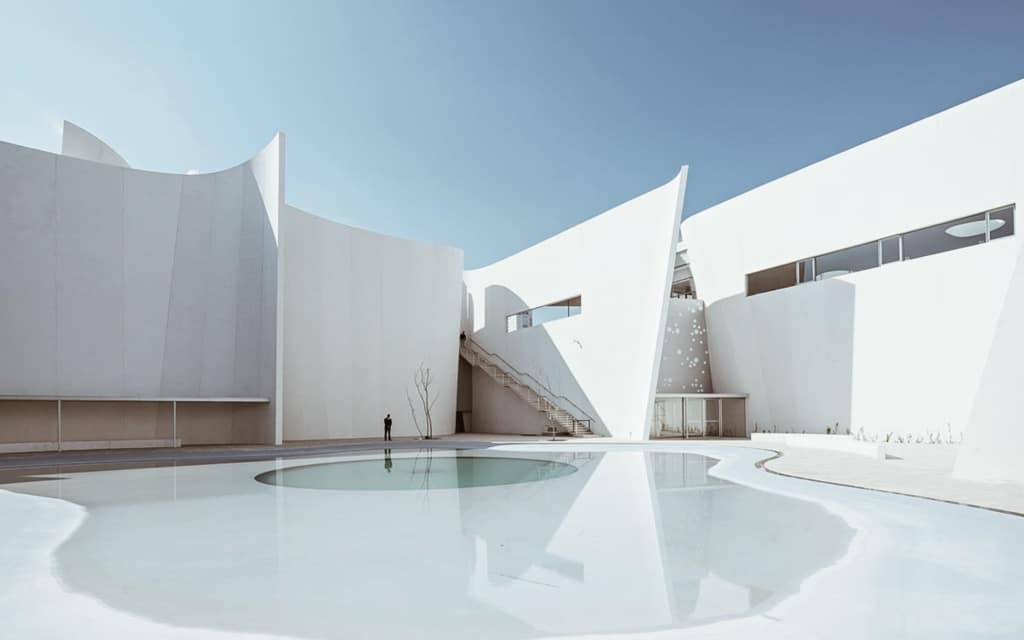 "A pristine white architectural design reflecting Toyo Ito's philosophy, featuring a sculpted building with a reflective water feature that highlights the harmony between nature and technology."