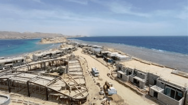 A panoramic view of the AMAALA construction site along the Red Sea, showcasing a series of modern structures under development. The buildings feature clean lines and contemporary design, blending seamlessly with the natural surroundings of sandy beaches and turquoise waters. Workers are actively engaged in construction activities, highlighting the progress being made on this ambitious project.