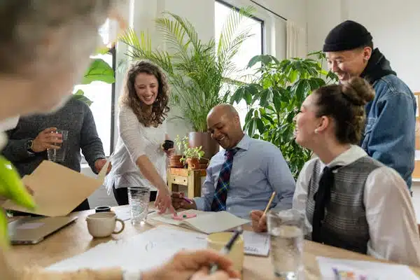 A group of people sitting around a work table, engaging in a collaborative discussion in a relaxed environment filled with green plants and natural light.