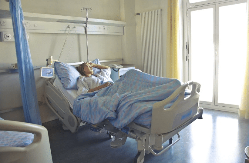 A hospital room with a comfortable bed, where a patient rests under soft-colored blankets, surrounded by natural light from large windows.