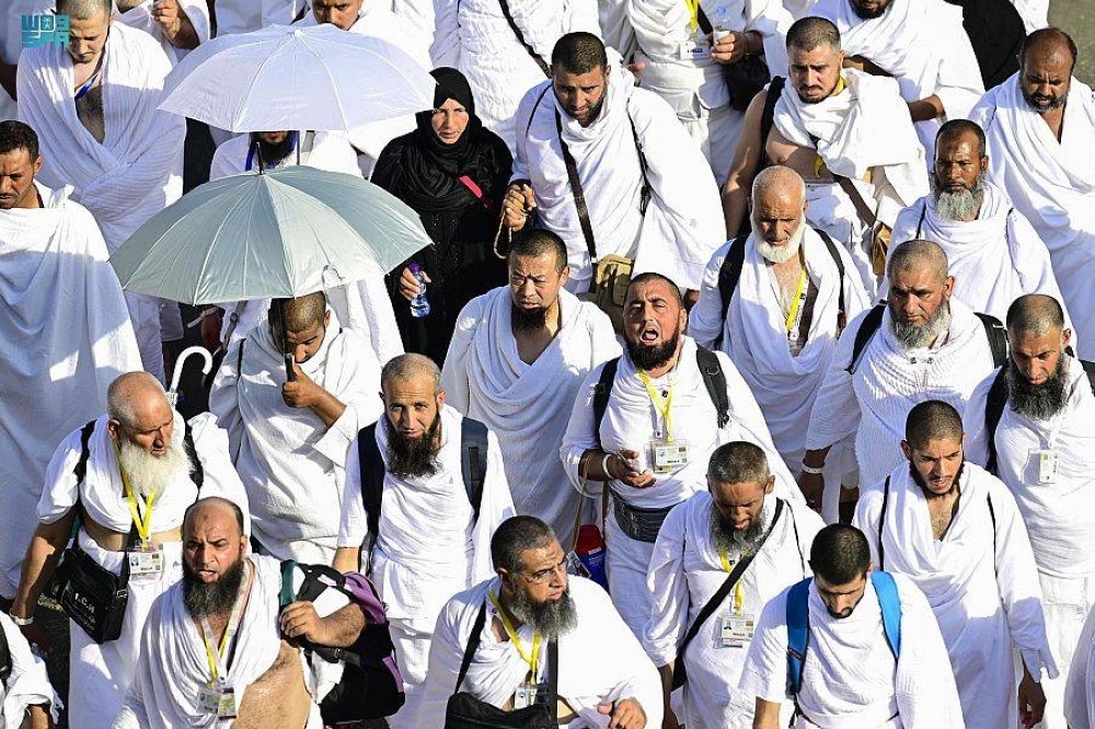 A large group of pilgrims walking together, all dressed in the distinctive white attire for Hajj and Umrah. Some pilgrims use umbrellas to shield themselves from the sun, while others carry bags and backpacks. Their faces reflect humility and spirituality.