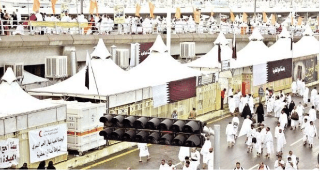 صورة تُظهر حشودًا من الحجاج يسيرون على طريق مخصص، جميعهم يرتدون الثياب البيضاء المميزة للحج والعمرة. في الخلفية، هناك خيم بيضاء كبيرة مرتبة بشكل منتظم، مع وجود لافتات وشعارات دينية وأعلام الدول المشاركة. الطريق مزدحم بالحجاج الذين يتحركون بسلاسة.