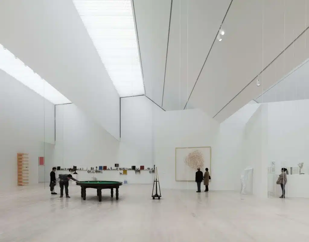 A spacious and minimalist art gallery with white walls, wooden floors, and large glass windows allowing natural light to flood the space.