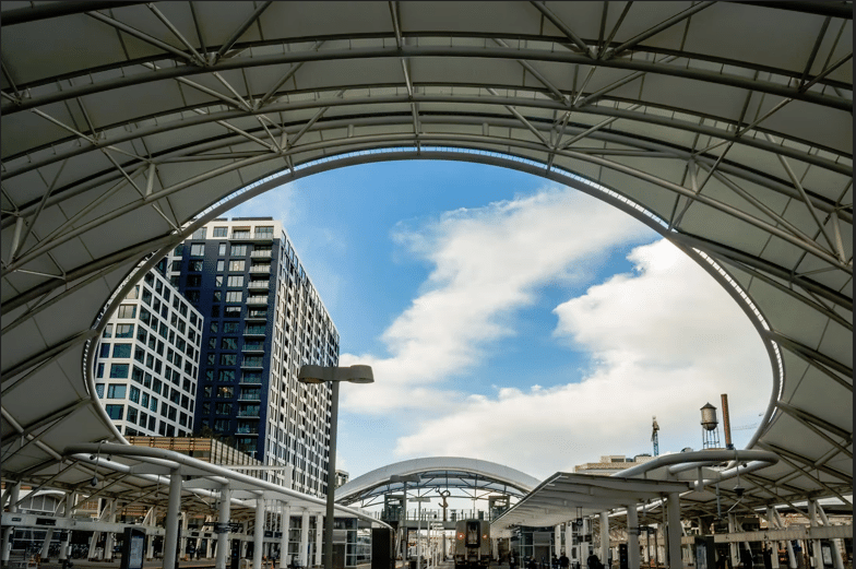 A tensile structure in the shape of an arch, combining cables and flexible membranes, showcasing a clear sky with white clouds and tall urban buildings in the background.