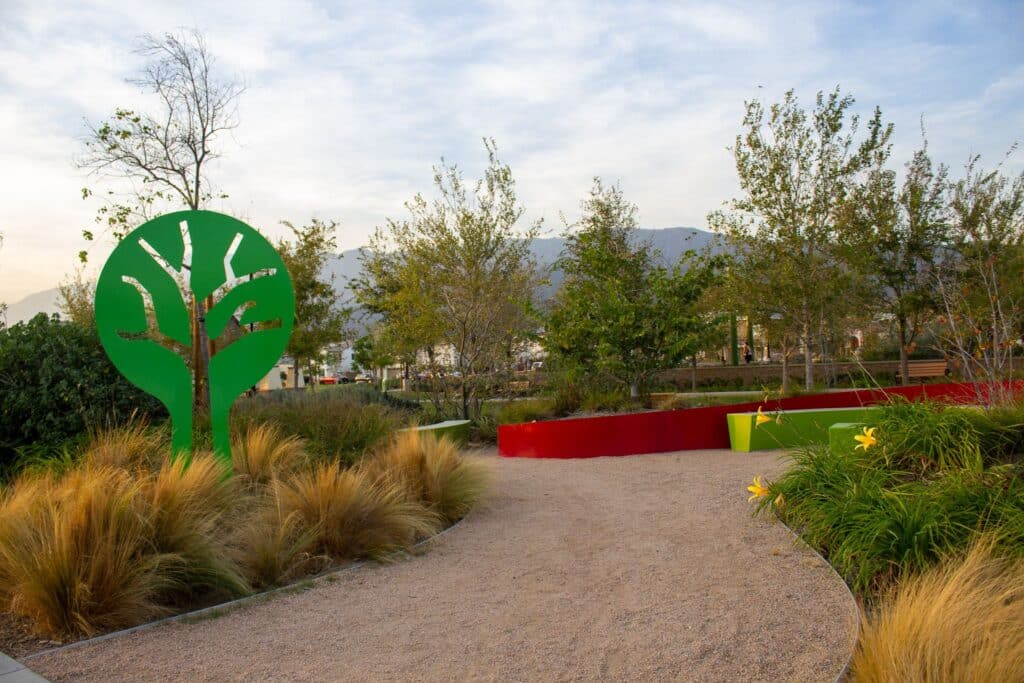 A park pathway featuring a large green tree sculpture on the left, surrounded by natural plants and vibrant yellow flowers, with decorative red and green walls in the background.
