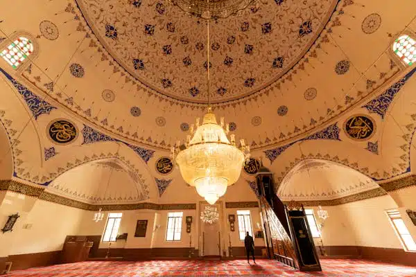 An ornate mosque dome decorated with intricate patterns and Quranic verses, featuring a grand chandelier that elegantly illuminates the space, with stained glass windows allowing natural light to enter.
